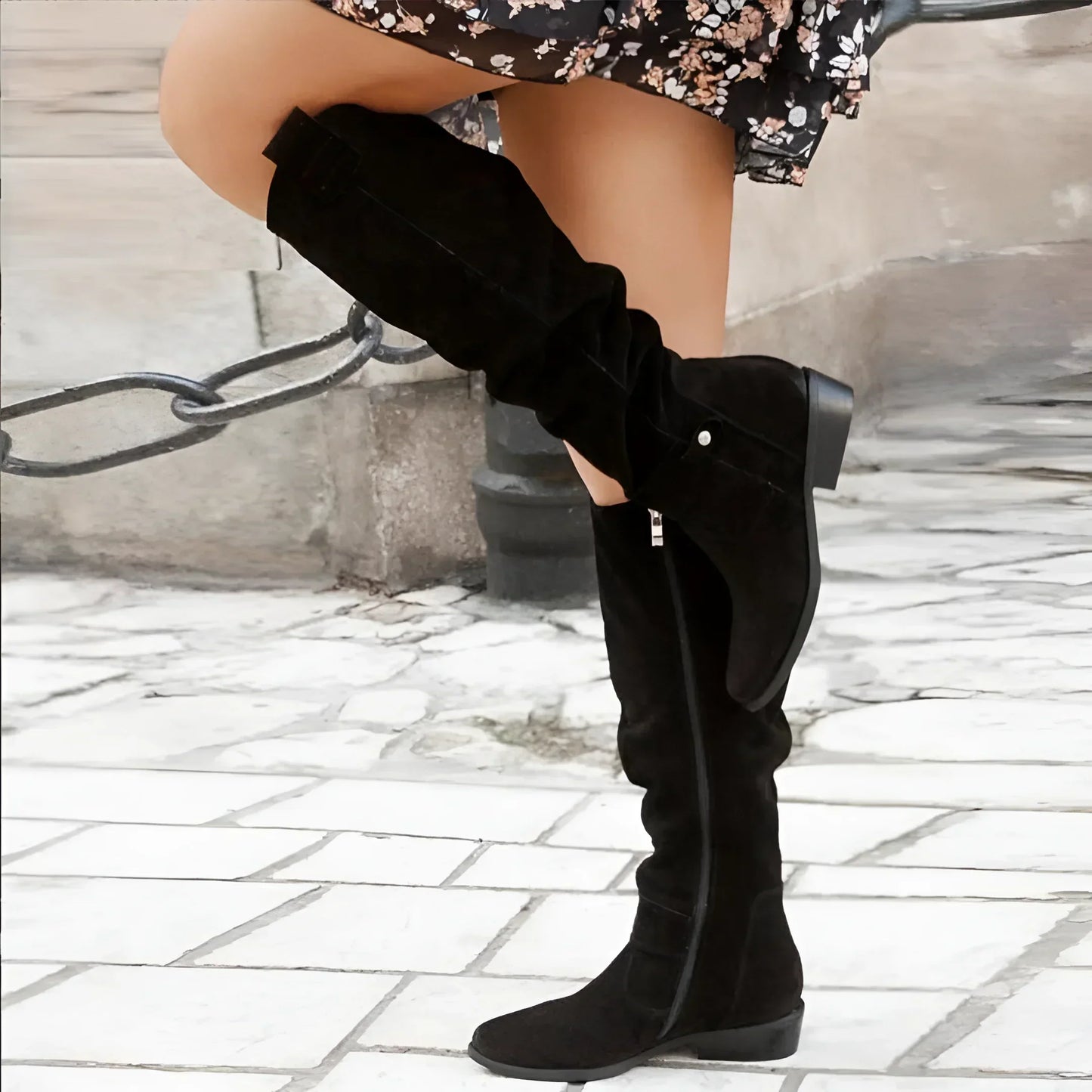 Black knee-high boots worn with a floral dress on a stone pavement background