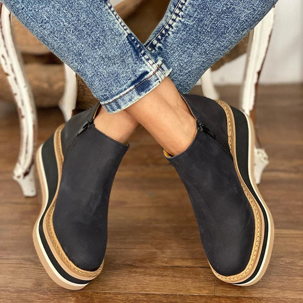 Black slip-on shoes with beige soles worn by a person sitting on a wooden floor