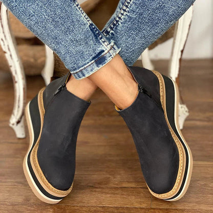Black slip-on shoes with beige soles worn by a person sitting on a wooden floor