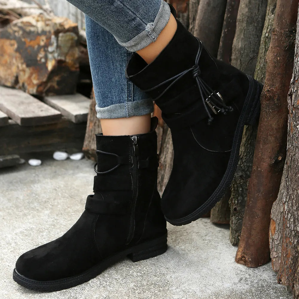 Black suede boots worn with rolled-up jeans against a rustic background