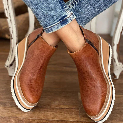 Brown leather boots with white soles worn by a person sitting on a wooden floor