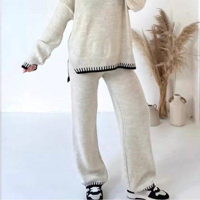 Person wearing a white sweater and pants with black trim, standing against a white wall with decorative elements